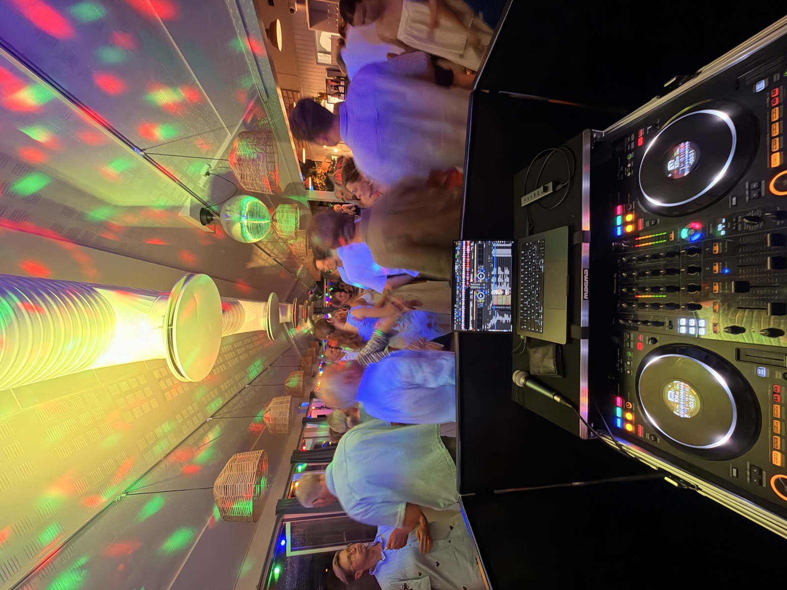 Fyldt dansegulv under discolys og en discokugle, set fra DJ-pulten